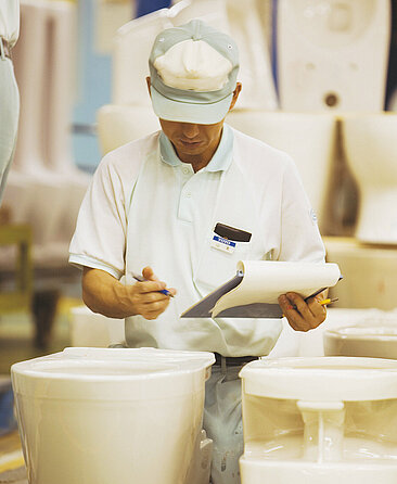 A man stands in front of some WC ceramics and looks at them closely. He is holding a clipboard with paper and a pen in his hands.